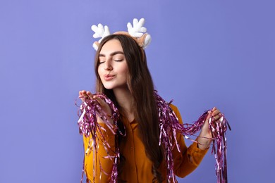 Photo of Young woman in reindeer headband with tinsel blowing kiss on purple background