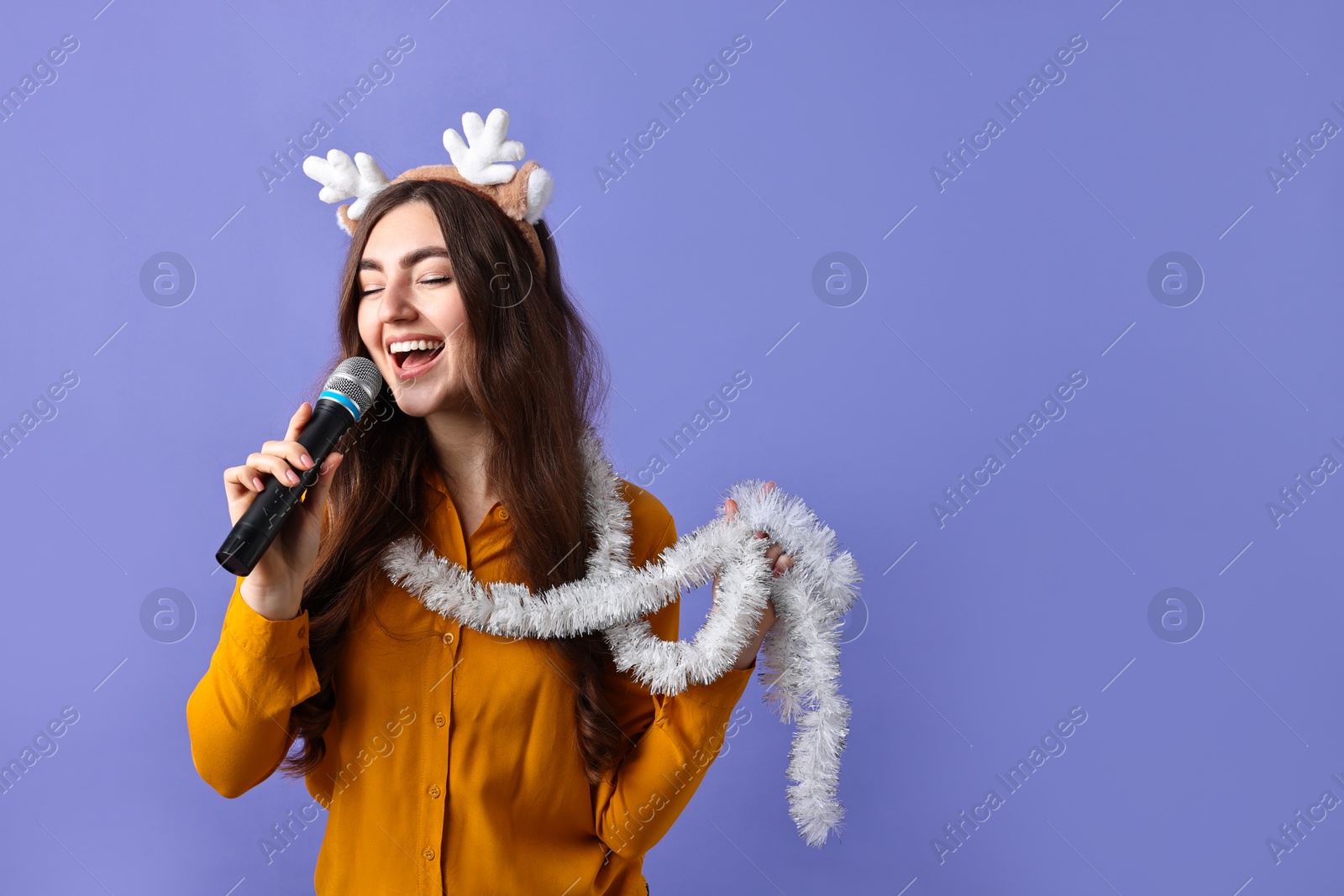 Photo of Young woman in reindeer headband with tinsel singing on purple background. Space for text