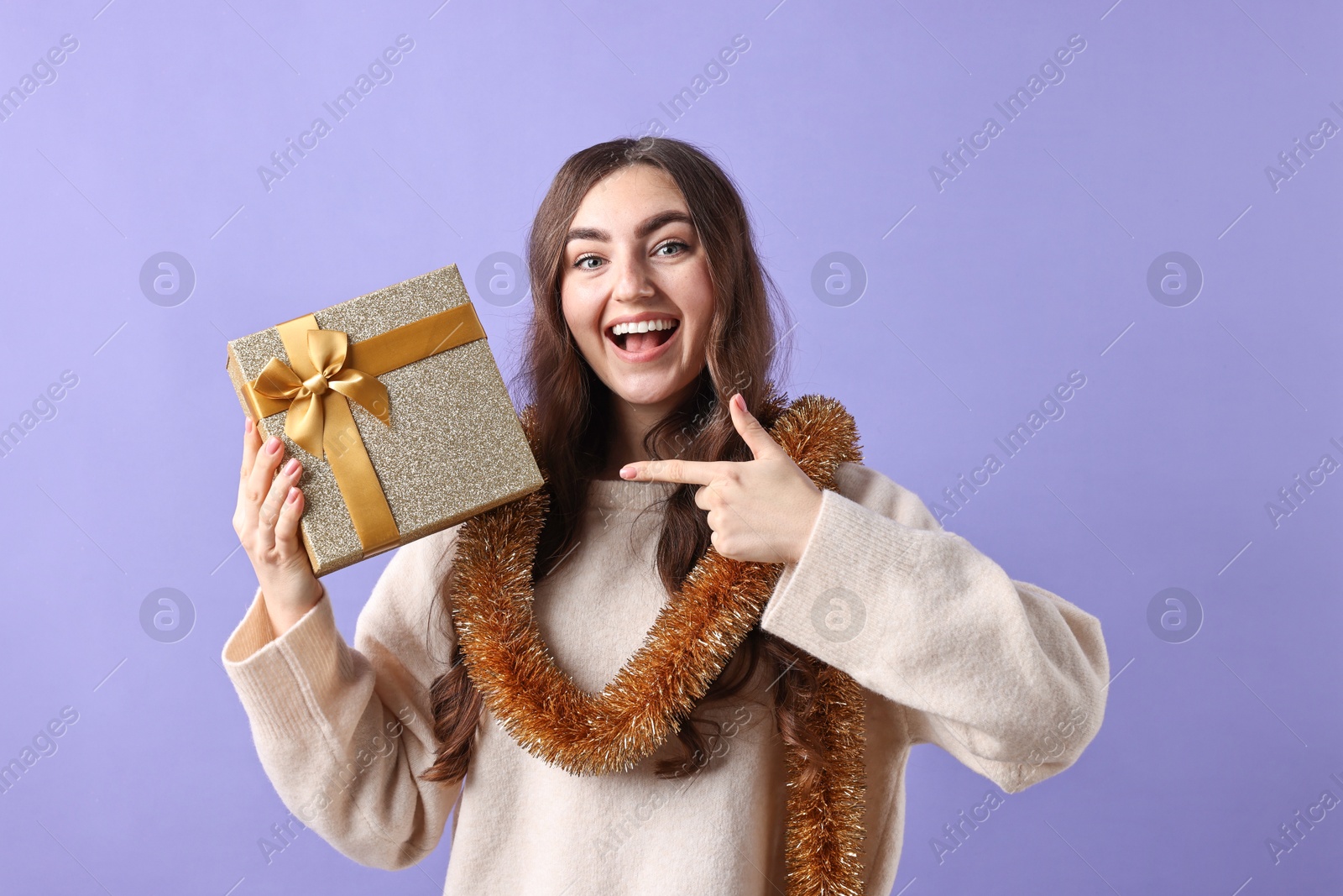 Photo of Happy young woman with tinsel and gift box on purple background