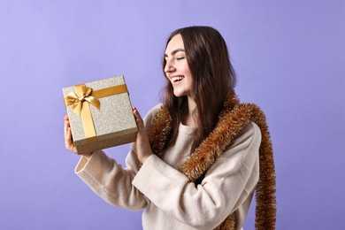 Photo of Happy young woman with tinsel and gift box on purple background