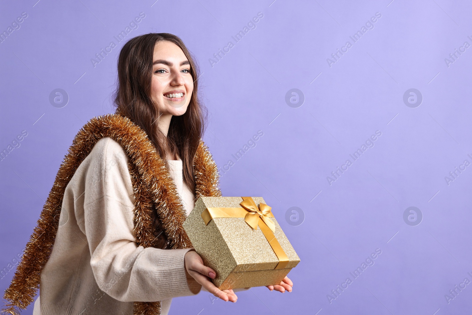 Photo of Happy young woman with tinsel and gift box on purple background. Space for text