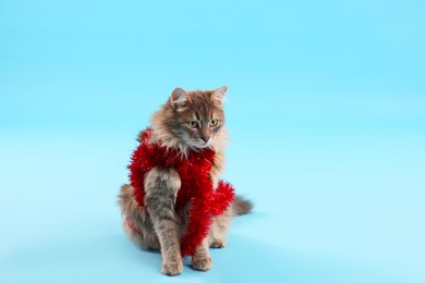 Photo of Cute cat with red Christmas tinsel on light blue background, space for text