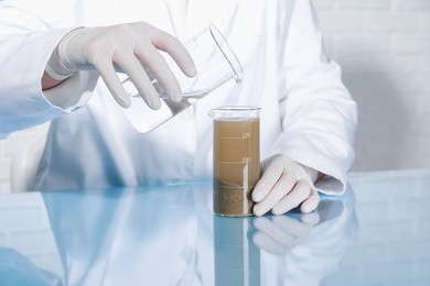 Photo of Laboratory testing. Scientist pouring liquid into beaker with soil sample at table indoors, closeup