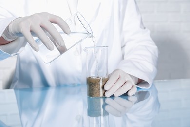 Photo of Laboratory testing. Scientist pouring liquid into beaker with soil sample at table indoors, closeup