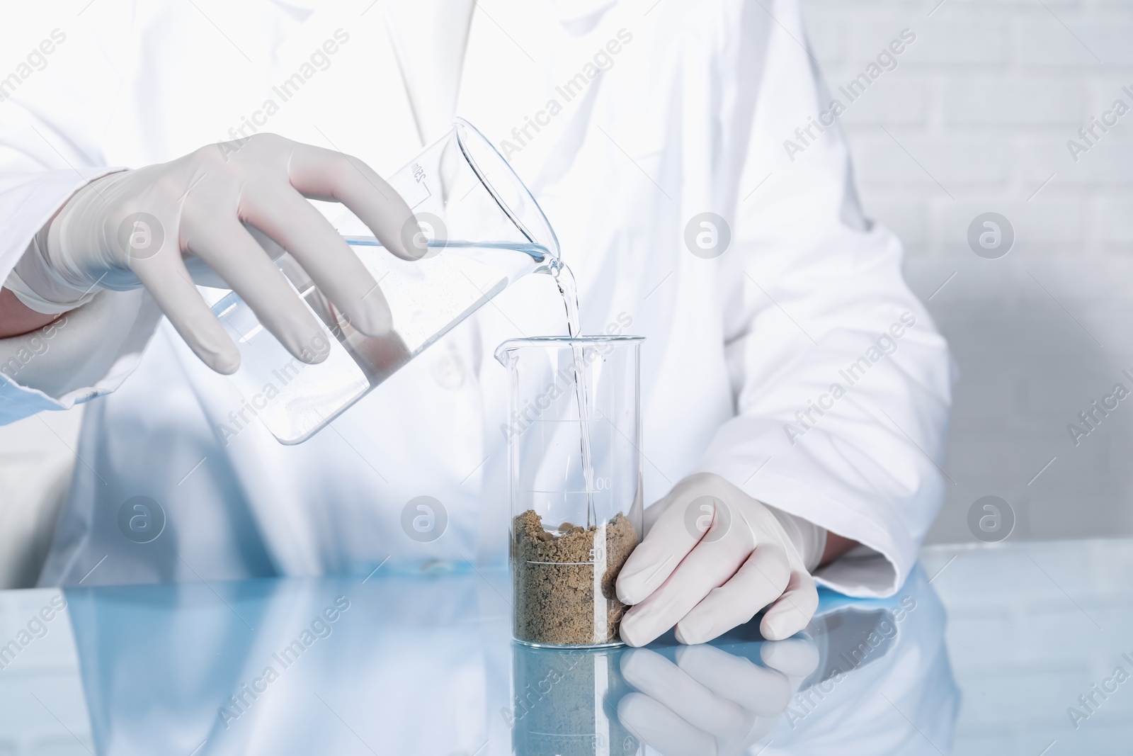 Photo of Laboratory testing. Scientist pouring liquid into beaker with soil sample at table indoors, closeup