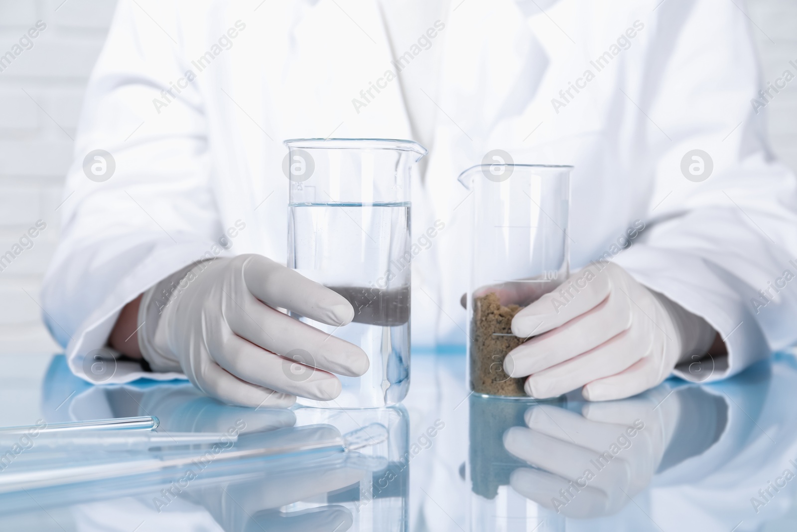 Photo of Laboratory testing. Scientist holding beakers with liquid and sand sample at table indoors, closeup