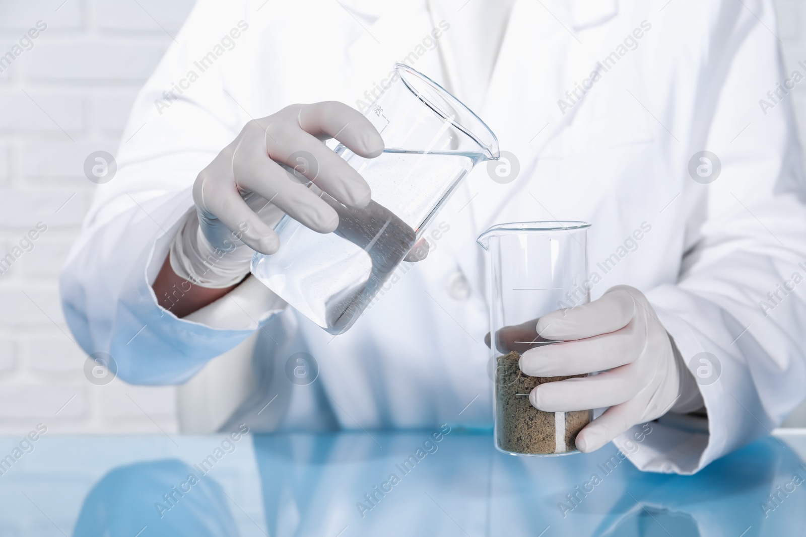 Photo of Laboratory testing. Scientist pouring liquid into beaker with soil sample at table indoors, closeup