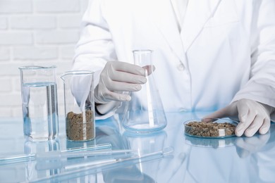 Photo of Laboratory testing. Scientist holding flask and petri dish with sand sample at table indoors, closeup