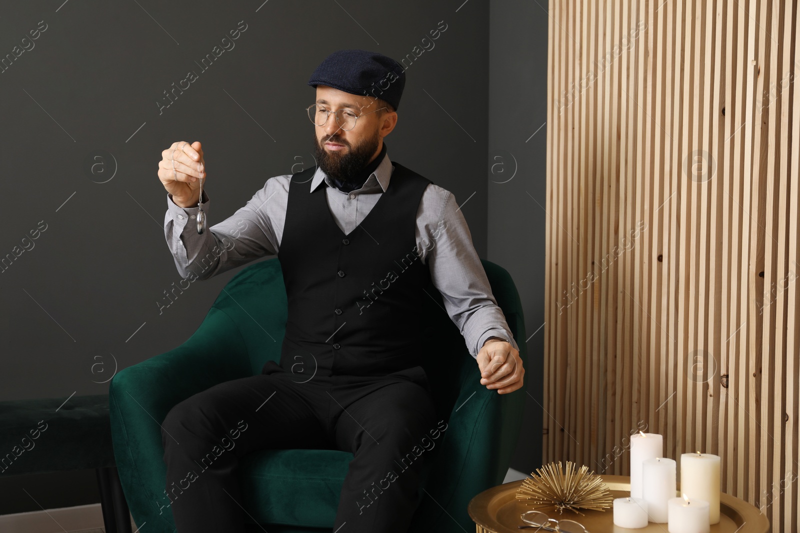 Photo of Hypnosis session. Man with vintage pocket watch in armchair near grey wall indoors