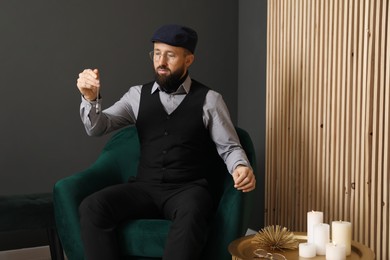 Photo of Hypnosis session. Man with vintage pocket watch in armchair near grey wall indoors