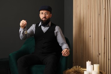 Photo of Hypnosis session. Man with vintage pocket watch in armchair near grey wall indoors