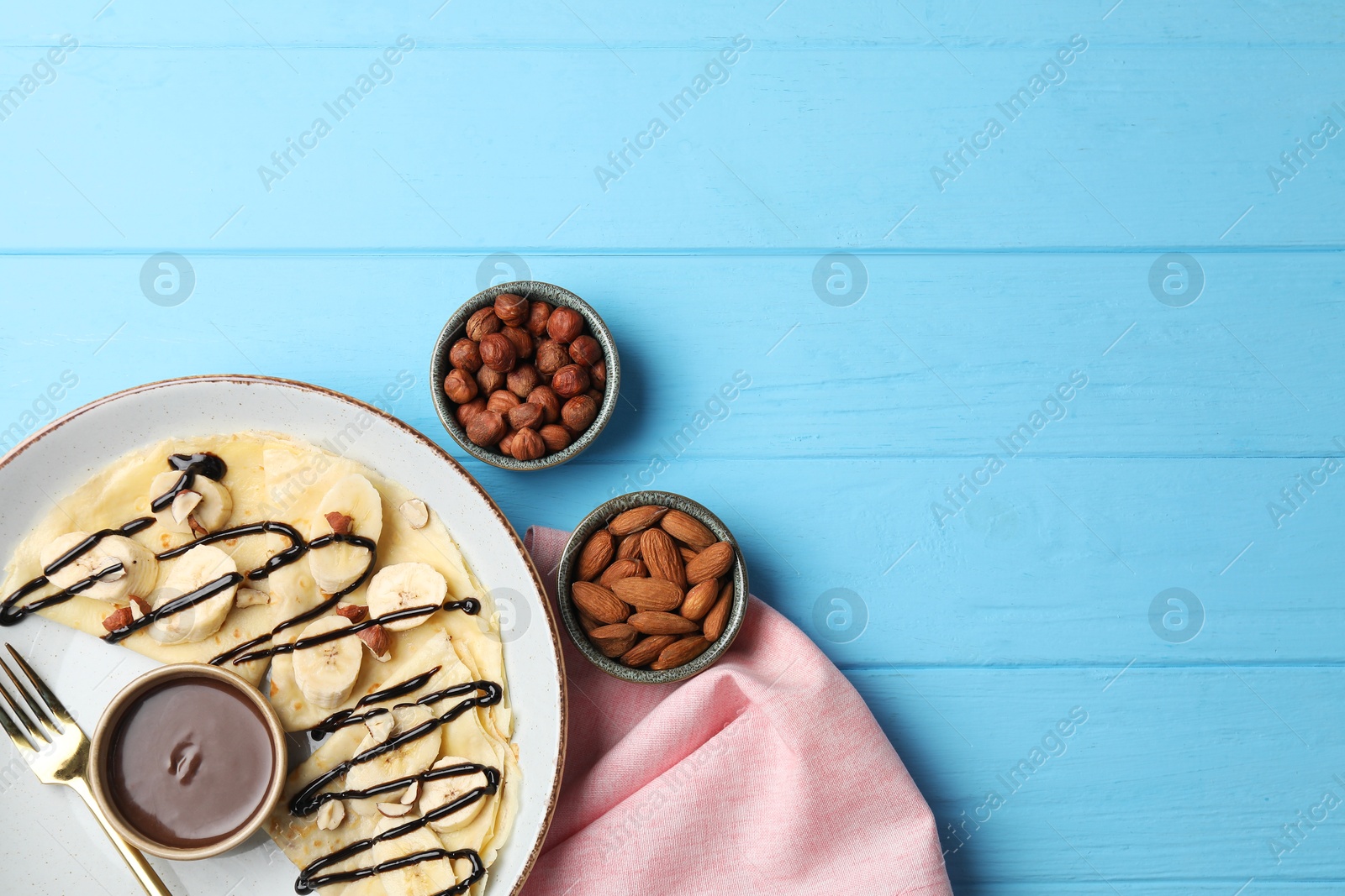 Photo of Delicious crepes with banana, nuts and chocolate sauce on light blue wooden table, flat lay. Space for text