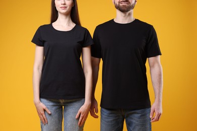 Photo of Woman and man wearing blank black t-shirts on orange background, closeup. Mockup for design