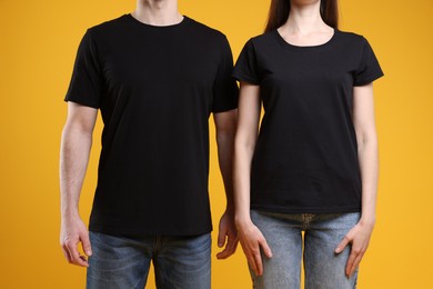 Photo of Woman and man wearing blank black t-shirts on orange background, closeup. Mockup for design