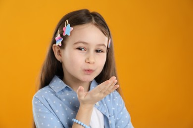 Photo of Little girl with beautiful hair clips blowing kiss on orange background, space for text