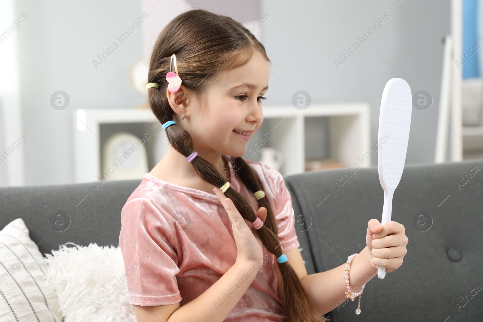 Photo of Little girl with beautiful hair accessories looking into mirror on sofa at home