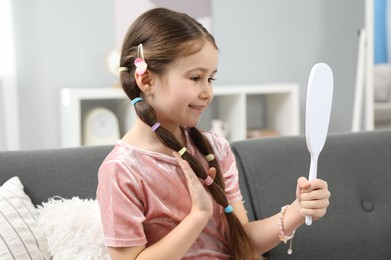 Photo of Little girl with beautiful hair accessories looking into mirror on sofa at home