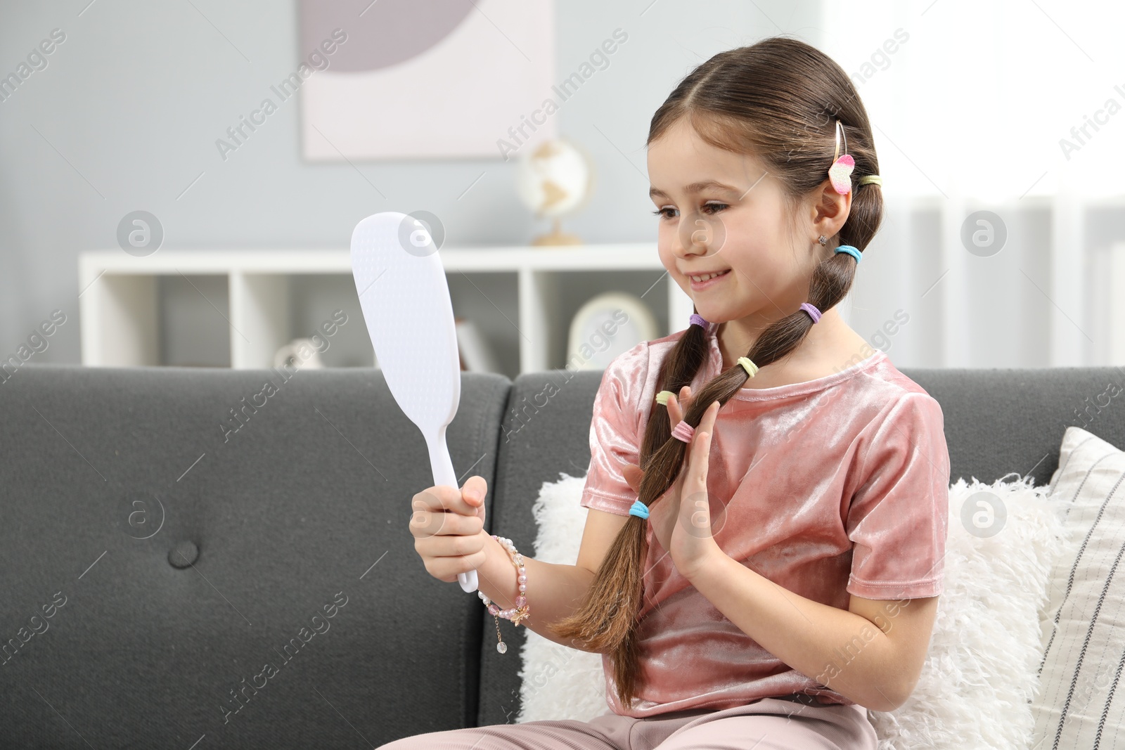 Photo of Little girl with beautiful hair accessories looking into mirror on sofa at home