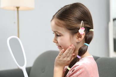 Photo of Little girl with beautiful hair accessories looking into mirror on sofa at home