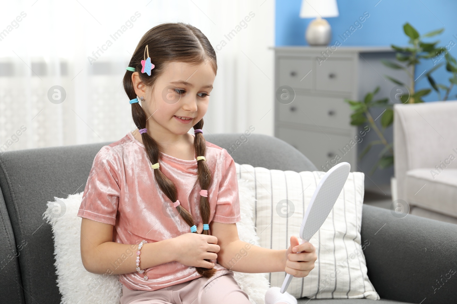 Photo of Little girl with beautiful hair accessories looking into mirror on sofa at home