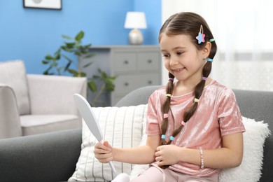 Photo of Little girl with beautiful hair accessories looking into mirror on sofa at home