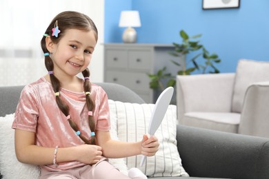 Photo of Little girl with beautiful hair accessories looking into mirror on sofa at home