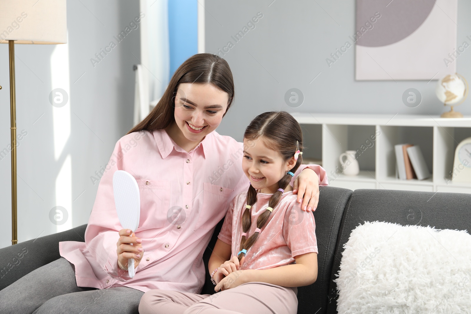 Photo of Cute little girl with hair accessories and her mom looking into mirror on sofa at home