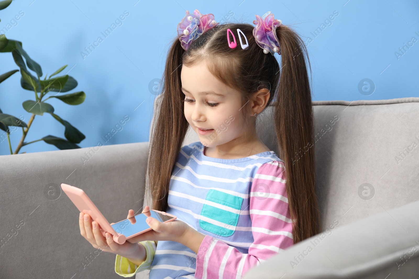 Photo of Little girl with beautiful hair accessories looking into pocket mirror in armchair at home