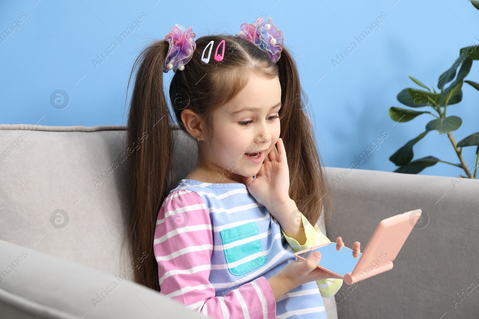 Photo of Little girl with beautiful hair accessories looking into pocket mirror in armchair at home