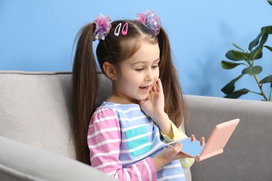 Photo of Little girl with beautiful hair accessories looking into pocket mirror in armchair at home