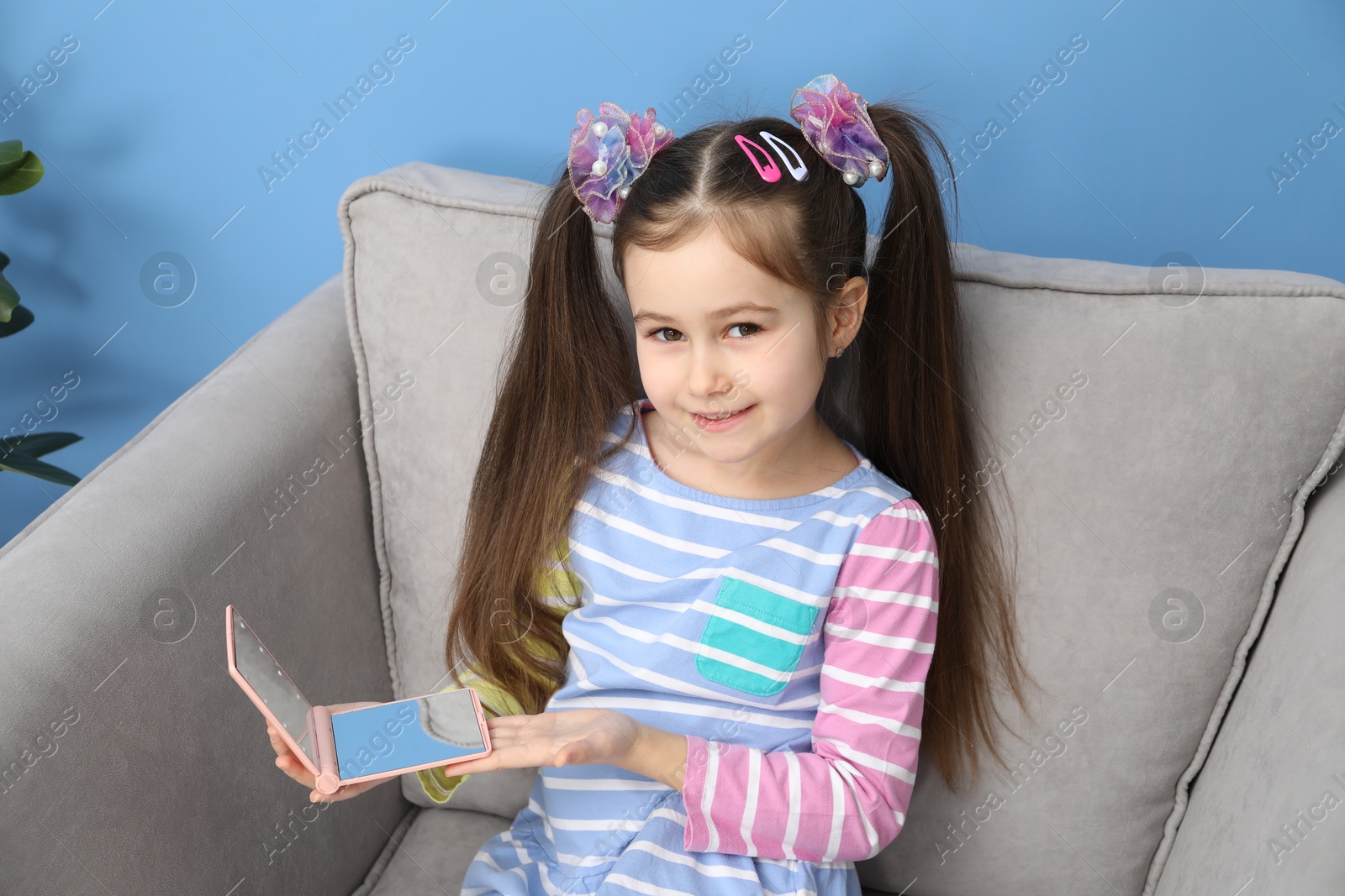 Photo of Little girl with beautiful hair accessories and pocket mirror in armchair at home