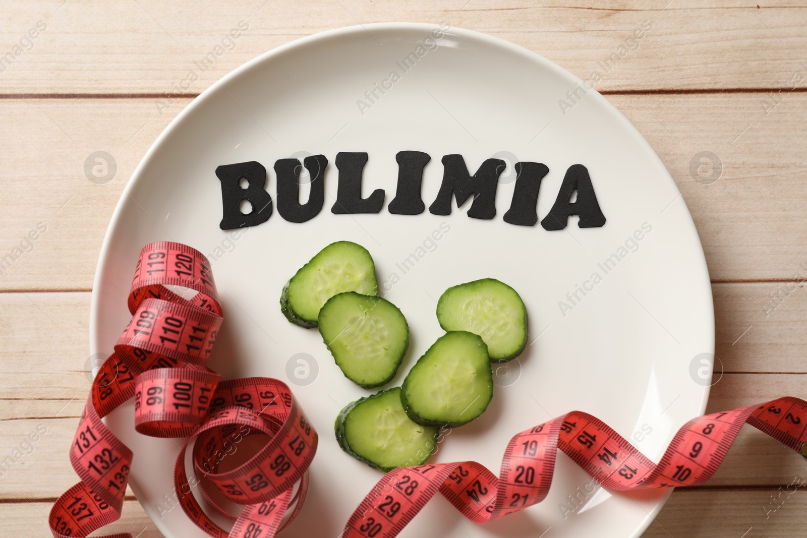 Photo of Plate with word Bulimia, cucumber slices and measuring tape on white wooden table, top view