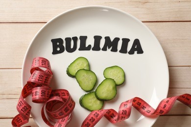 Photo of Plate with word Bulimia, cucumber slices and measuring tape on white wooden table, top view