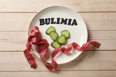 Photo of Plate with word Bulimia, cucumber slices and measuring tape on white wooden table, top view