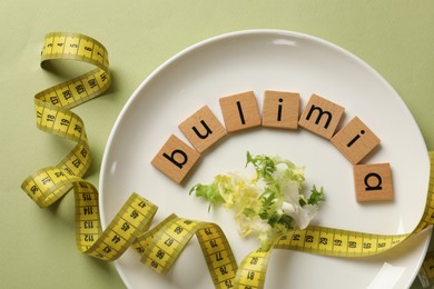 Photo of Plate with word Bulimia, lettuce and measuring tape on olive background, top view