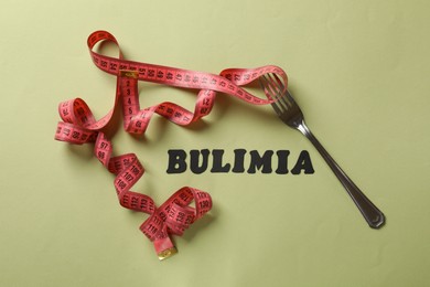 Photo of Word Bulimia, measuring tape and fork on olive background, top view