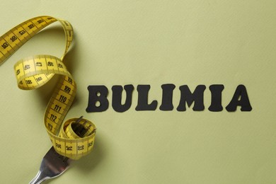 Photo of Word Bulimia, measuring tape and fork on olive background, top view