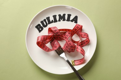 Photo of Plate with word Bulimia, measuring tape and fork on olive background, top view