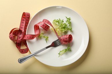 Photo of Bulimia. Plate with lettuce, fork and measuring tape on beige background, top view