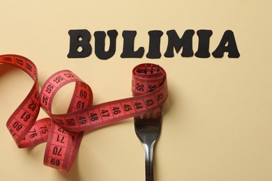 Photo of Word Bulimia, fork and measuring tape on beige background, top view