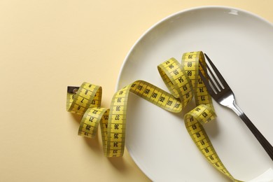 Photo of Bulimia. Plate with fork and measuring tape on beige background, top view