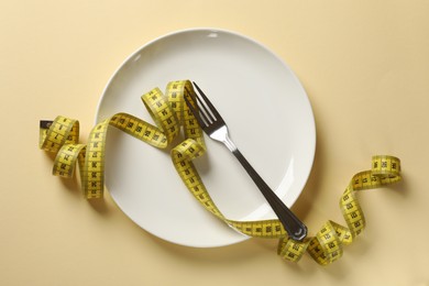 Photo of Bulimia. Plate with fork and measuring tape on beige background, top view