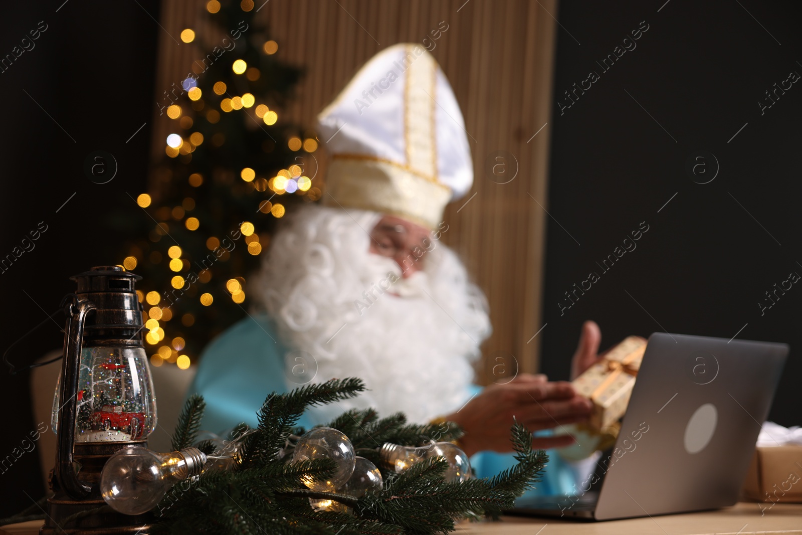 Photo of Saint Nicholas with gift and laptop in room decorated for Christmas, selective focus