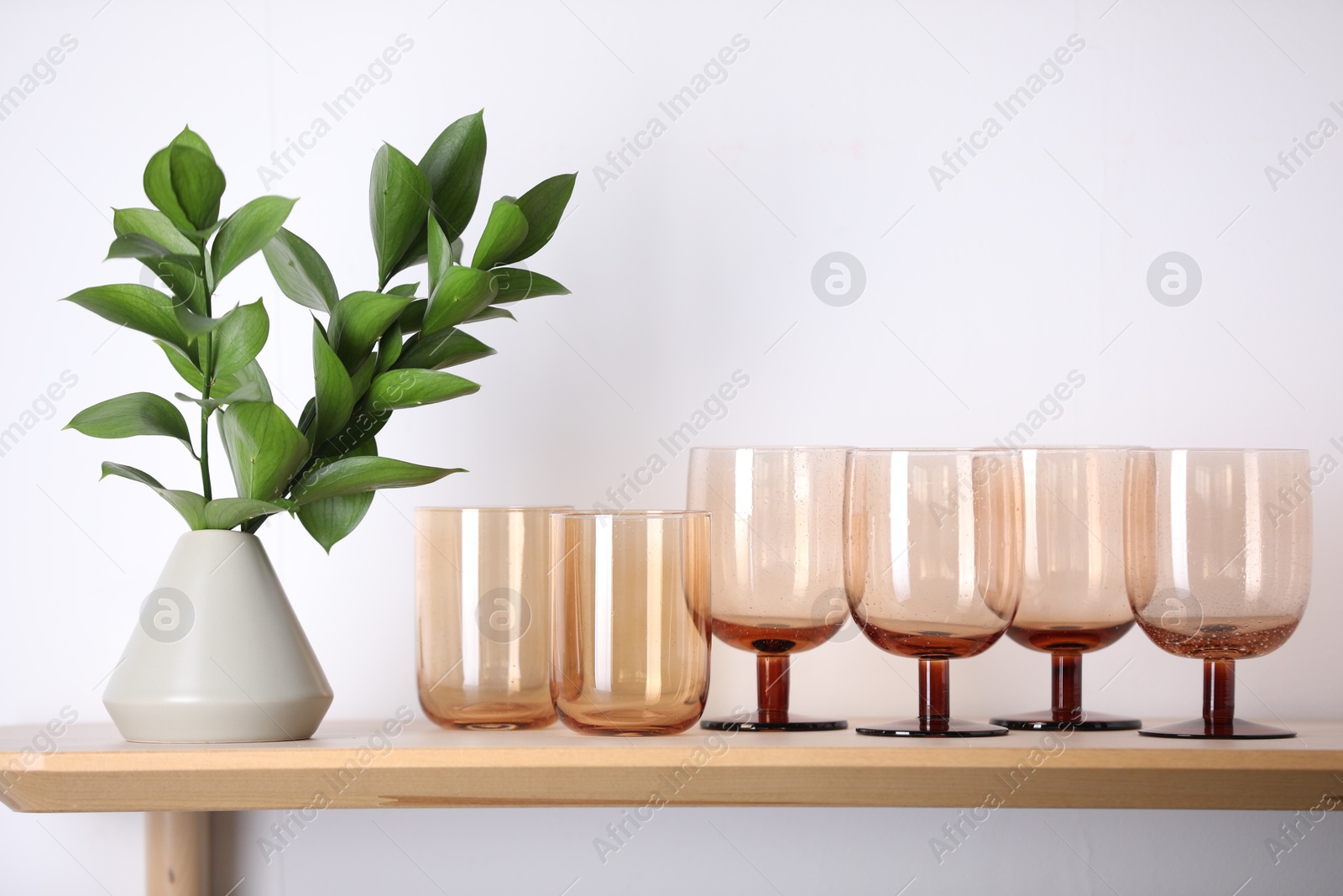 Photo of Empty clean glasses and green branches on wooden shelf near white wall indoors