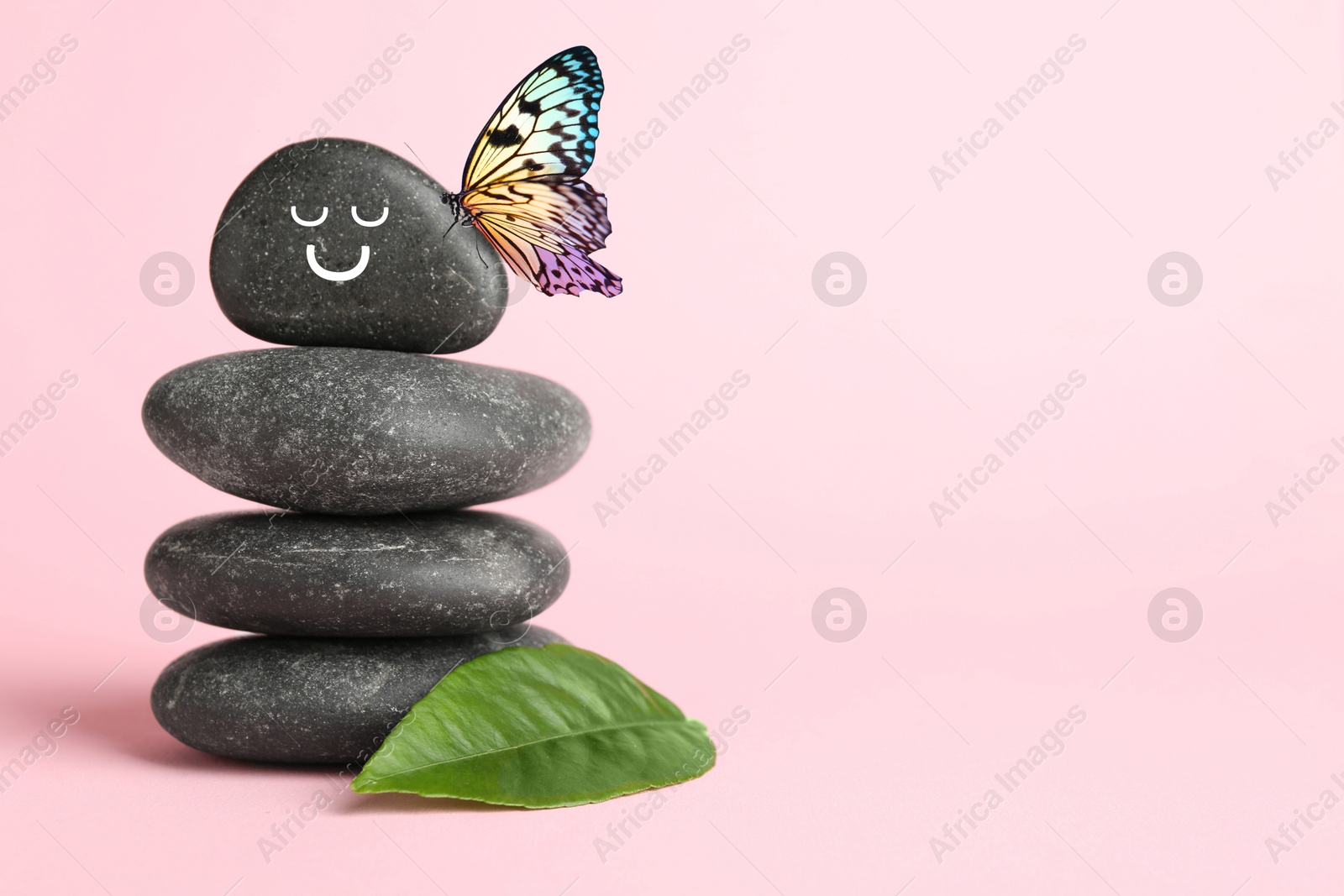 Image of Butterfly on stone with smiling face, stack of pebbles and green leaf on pink background, space for text. Harmony