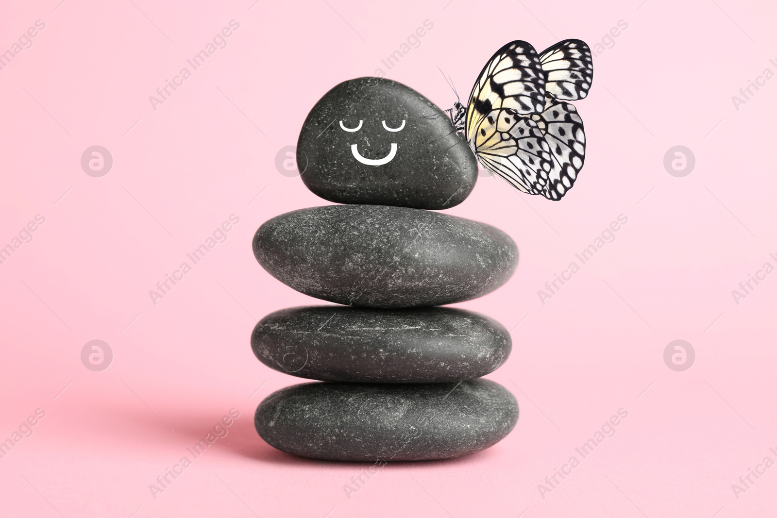 Image of Butterfly on stone with smiling face, stack of pebbles on pink background. Harmony