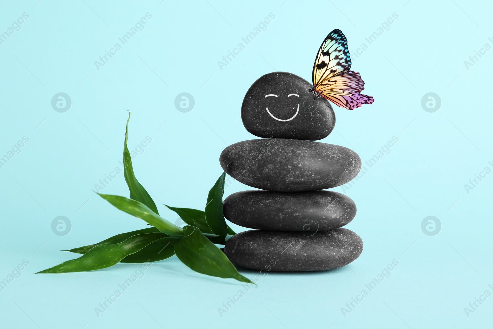 Image of Butterfly on stone with smiling face, stack of pebbles and green branch on light turquoise background. Harmony