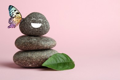 Image of Butterfly on stone with smiling face, stack of pebbles and green leaf on pink background, space for text. Harmony