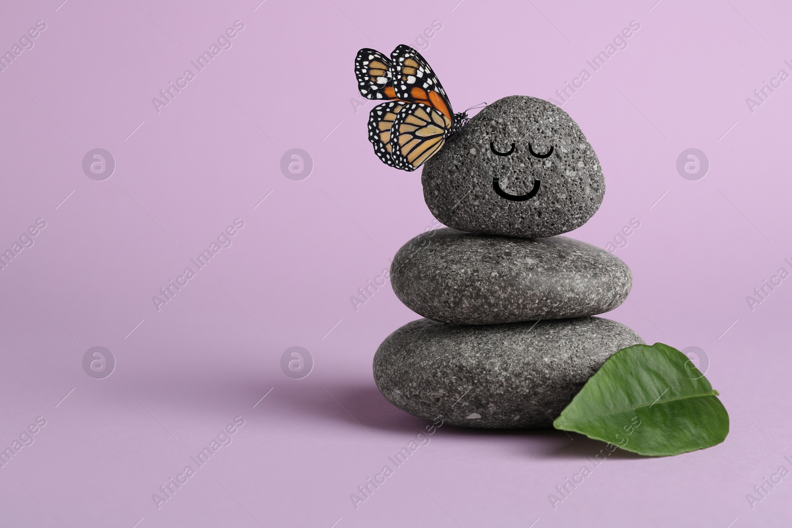 Image of Butterfly on stone with smiling face, stack of pebbles and green leaf on light violet background, space for text. Harmony