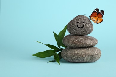 Image of Butterfly on stone with smiling face, stack of pebbles and green branch on light blue background, space for text. Harmony